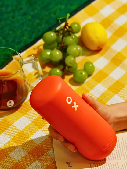 Person holding an orange portable grinding device on a yellow checkered tablecloth with fruits in the background