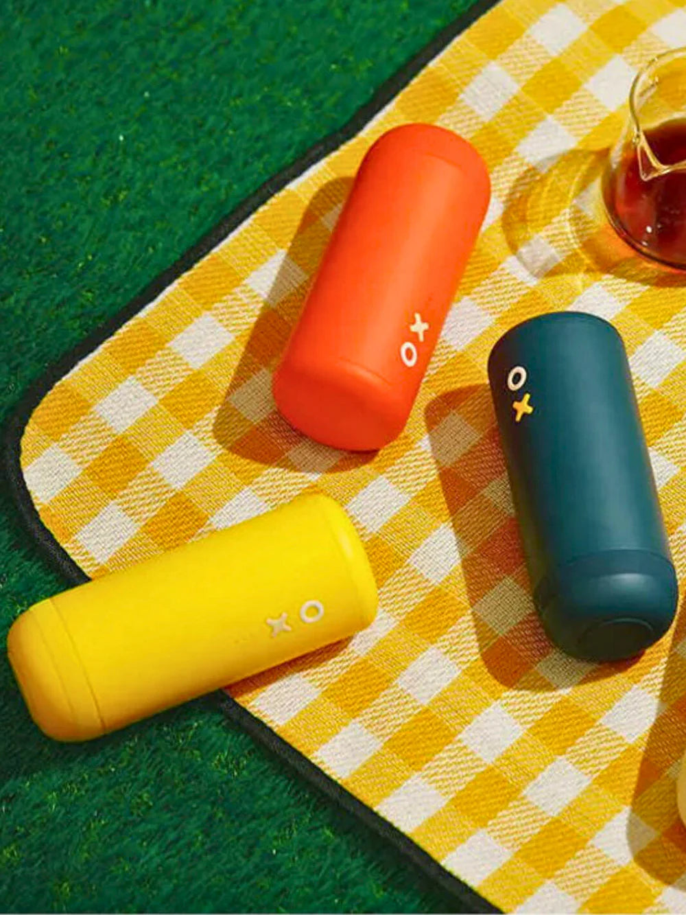Three colorful coffee grinders on a yellow checkered tablecloth with a glass of tea in the background.