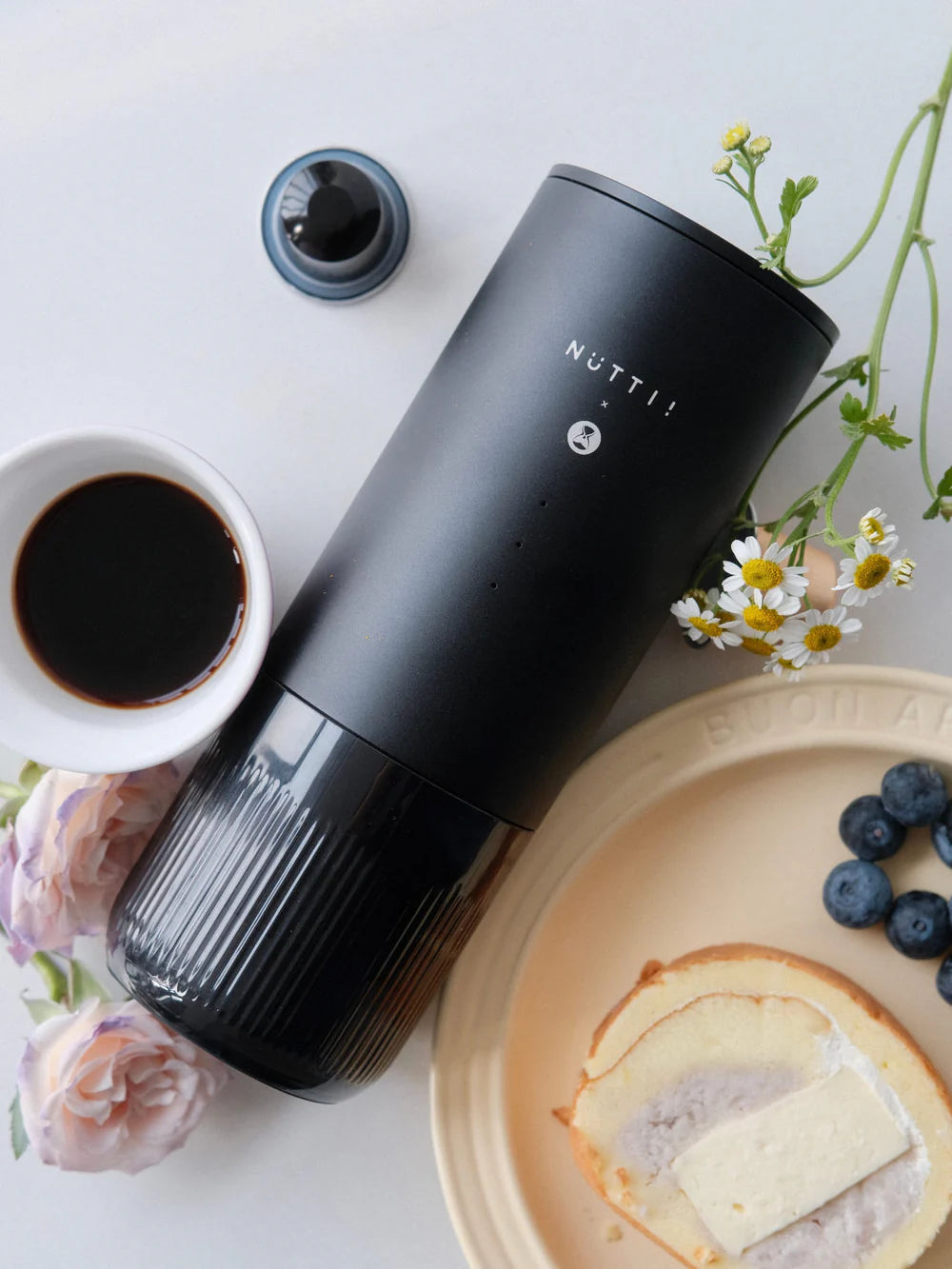 Black portable espresso machine with a cup of coffee, plate of cake, and flowers on a white surface