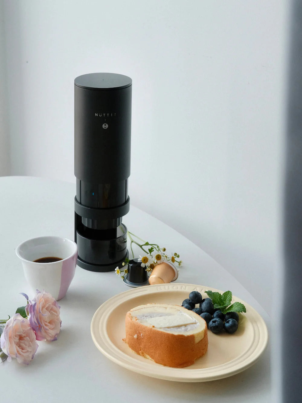 Black coffee maker on a white table with a cup of coffee, plate of bread and berries, and flowers.