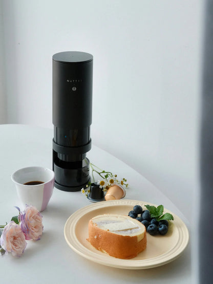 Black coffee maker on a white table with a cup of coffee, plate of bread and berries, and flowers.