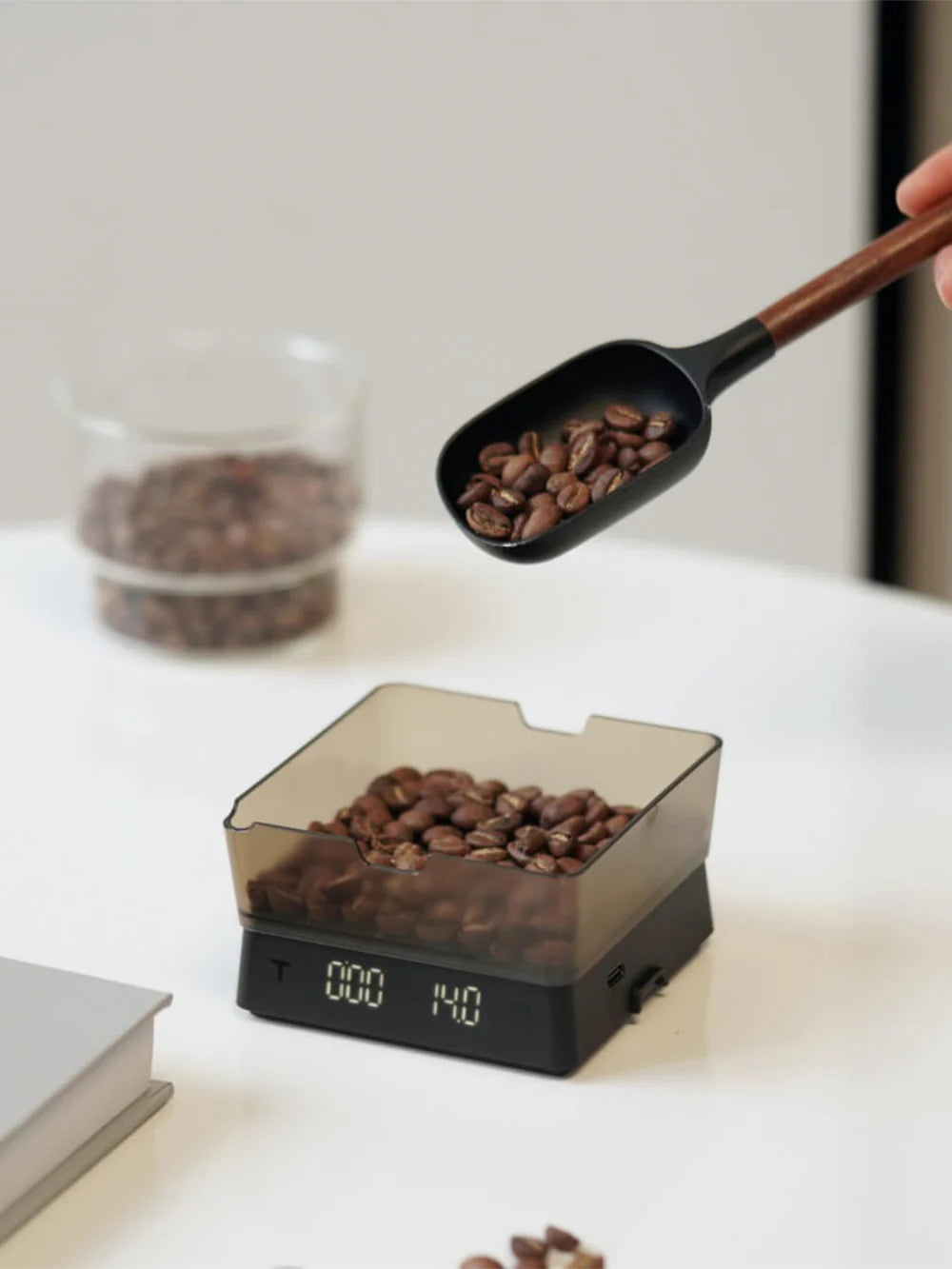 Coffee scoop filling a digital coffee scale with coffee beans on a white surface.