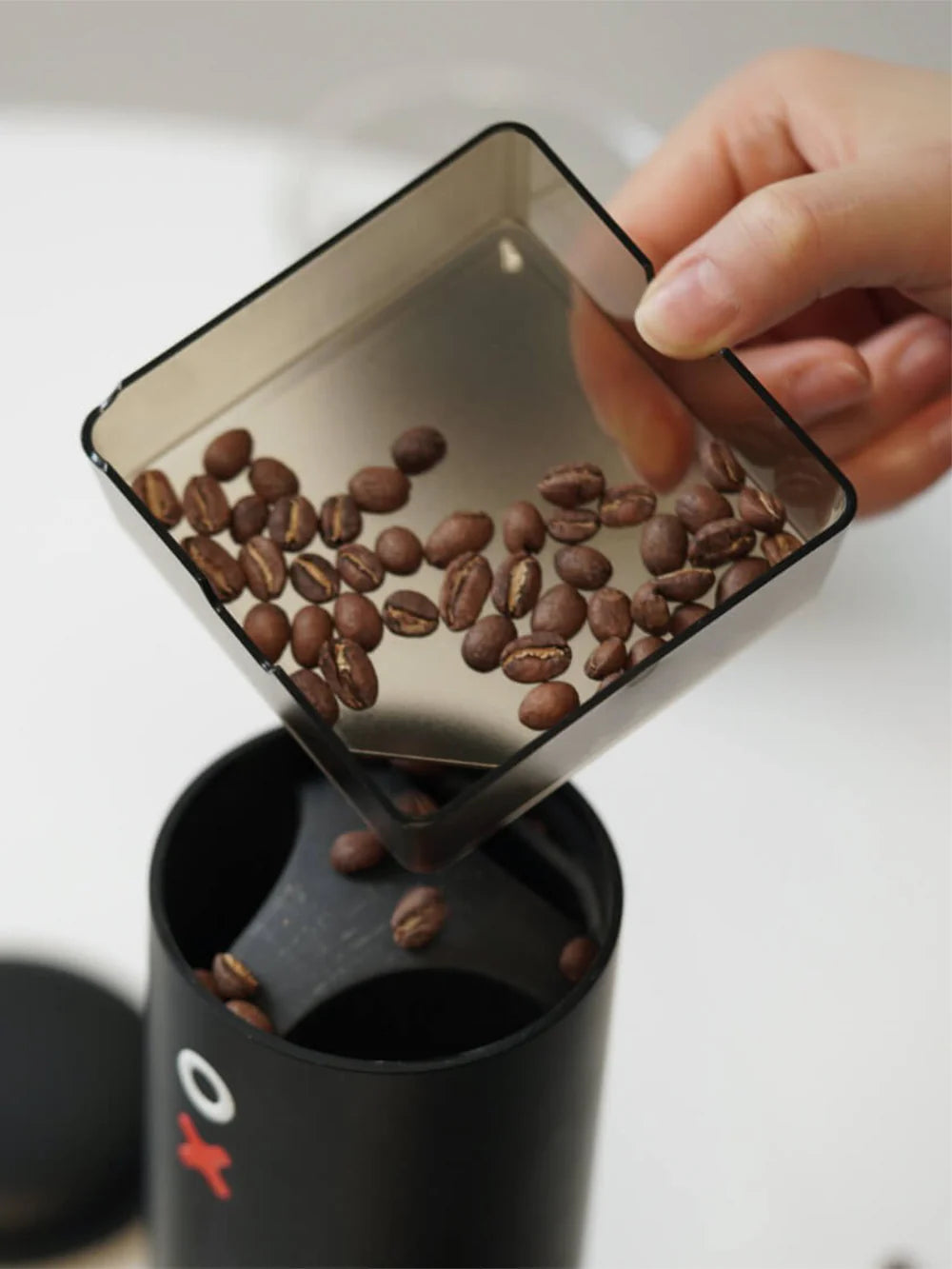Hand pouring coffee beans from a transparent container into a black coffee grinder.
