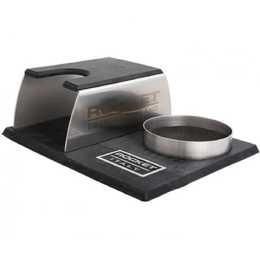 A Rocket Espresso Milano espresso tamping station featuring a brushed stainless steel body with black rubberized elements and a matching metal tamper holder on a black base.