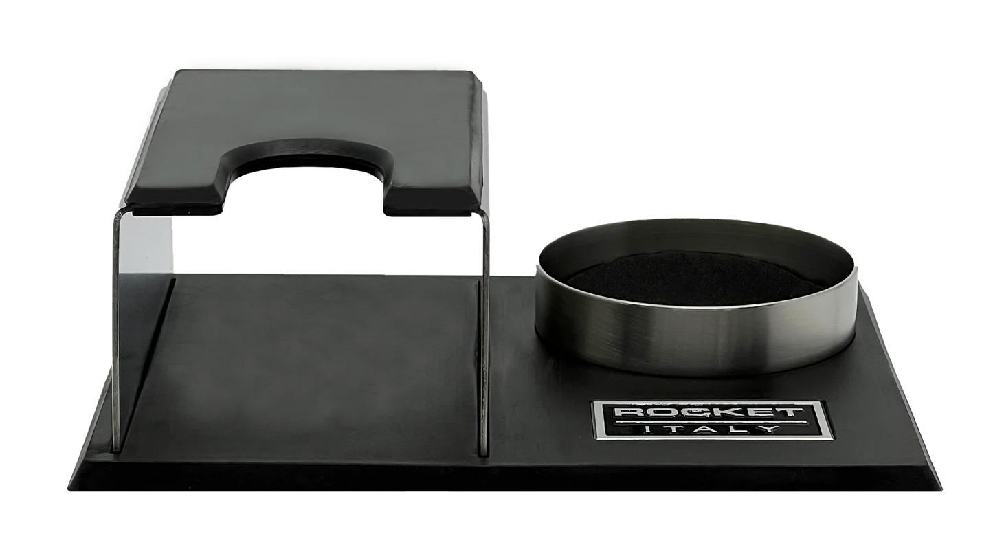 A Rocket Espresso Milano espresso tamping station featuring a brushed stainless steel body with black rubberized elements and a matching metal tamper holder on a black base. Front view.
