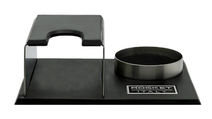 A Rocket Espresso Milano espresso tamping station featuring a brushed stainless steel body with black rubberized elements and a matching metal tamper holder on a black base. Front view.