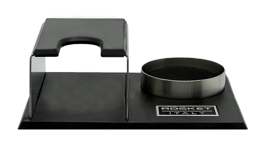 A Rocket Espresso Milano espresso tamping station featuring a brushed stainless steel body with black rubberized elements and a matching metal tamper holder on a black base. Front view.