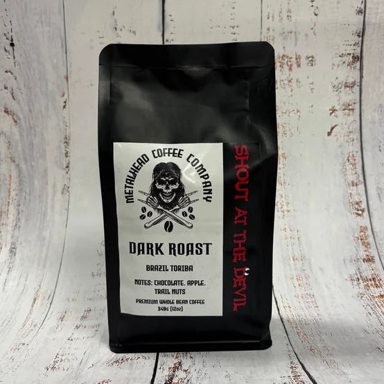 A black coffee bag labeled 'Metalhead Coffee Company' with a white front label and red vertical text "Shout at the devil", standing on a distressed white wood surface.