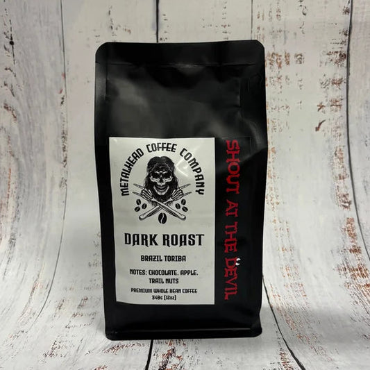 A black coffee bag labeled 'Metalhead Coffee Company' with a white front label and red vertical text "Shout at the devil", standing on a distressed white wood surface.