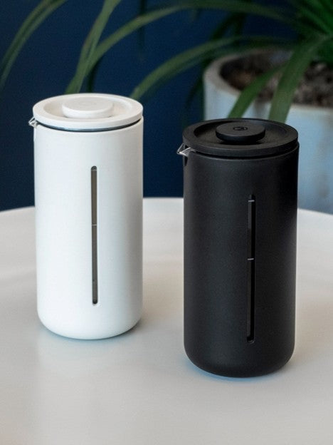White and black travel mugs on a light surface with a plant in the background