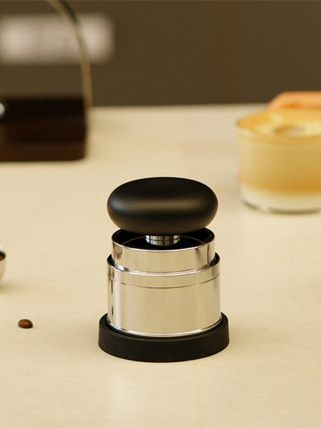 Coffee tamp on a table with coffee beans and a blurred beverage in the background.