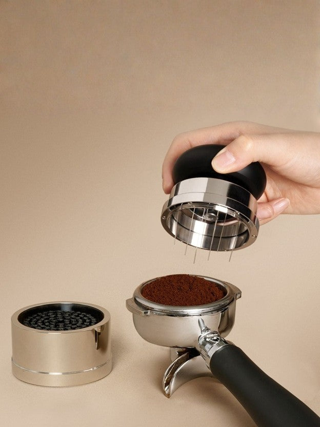 Hand holding a needle distribution tool near a portafilter with ground espresso beans on a beige background