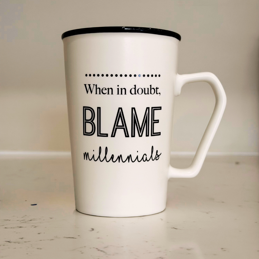 White mug with black text 'When in doubt, blame millennials' on a light wooden surface.