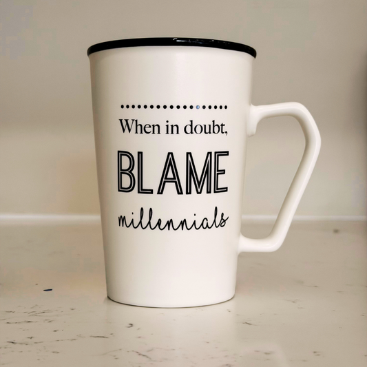 White mug with black text 'When in doubt, blame millennials' on a light wooden surface.