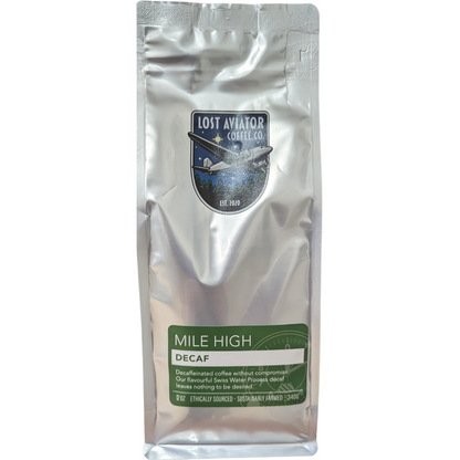 A package of Lost Aviator Mile High Decaf coffee with the brand's logo and product name visible. The packaging is silver in color with green text and a green geometric design.