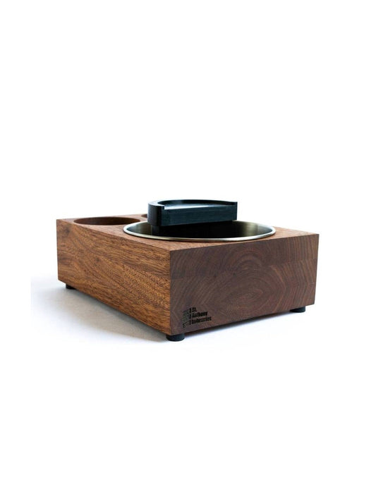 A brown wooden espresso knock box and tamp station with a stainless steel knock box reservoir and a black plastic tamping stand.