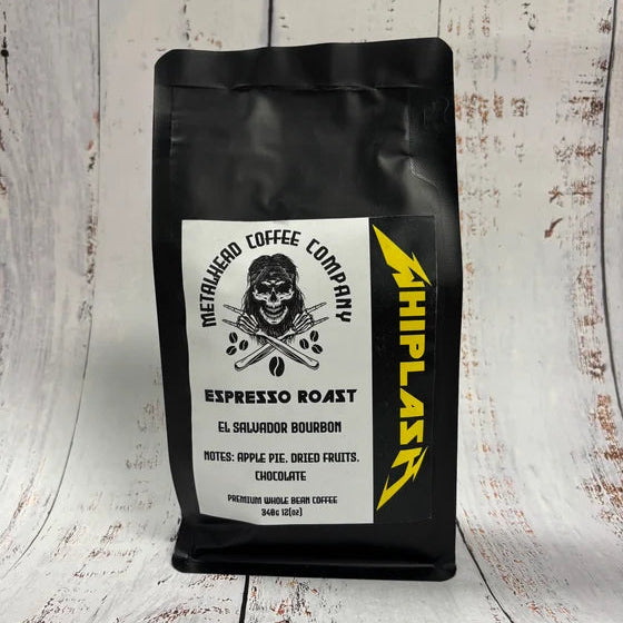 A black bag of MetaRuedo Coffee Company Espresso Roast, featuring a white label with a skull graphic and a yellow 'WHIPLAST' logo, set against a rustic white-painted wood background.