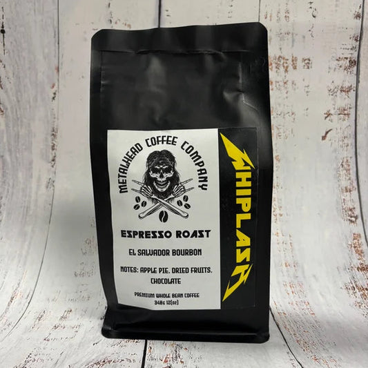 A black bag of MetaRuedo Coffee Company Espresso Roast, featuring a white label with a skull graphic and a yellow 'WHIPLAST' logo, set against a rustic white-painted wood background.