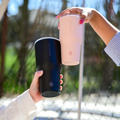 Two hands holding insulated travel mugs outdoors with a blurred natural background