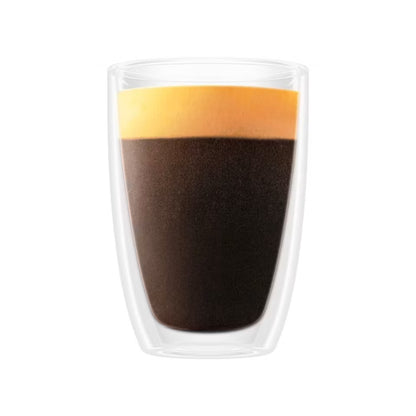 A clear double-walled glass cup containing dark coffee with a layer of orange-yellow foam on top, centered on a plain white background.