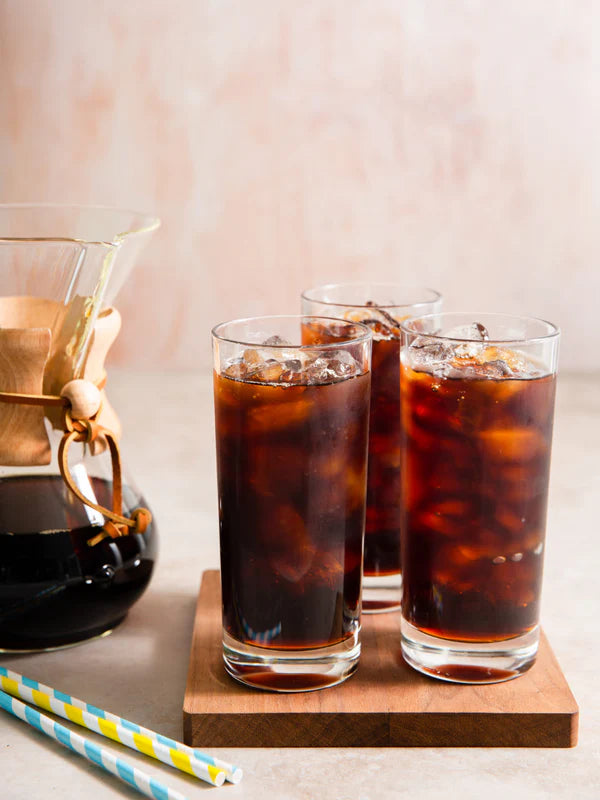 Three glasses of iced coffee on a wooden coaster with a coffee chemex in the background.