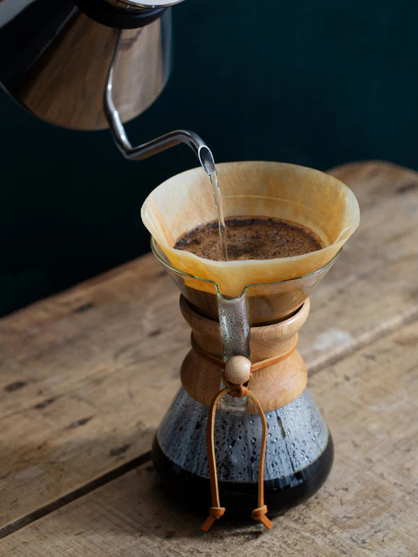 Coffee being poured into a Chemex coffee maker on a wooden surface.
