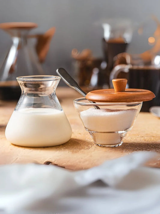 A clear glass milk carafe filled with milk and a small glass bowl with a wooden lid, sugar inside, and a spoon, set on a wooden table with a blurred kitchen background.