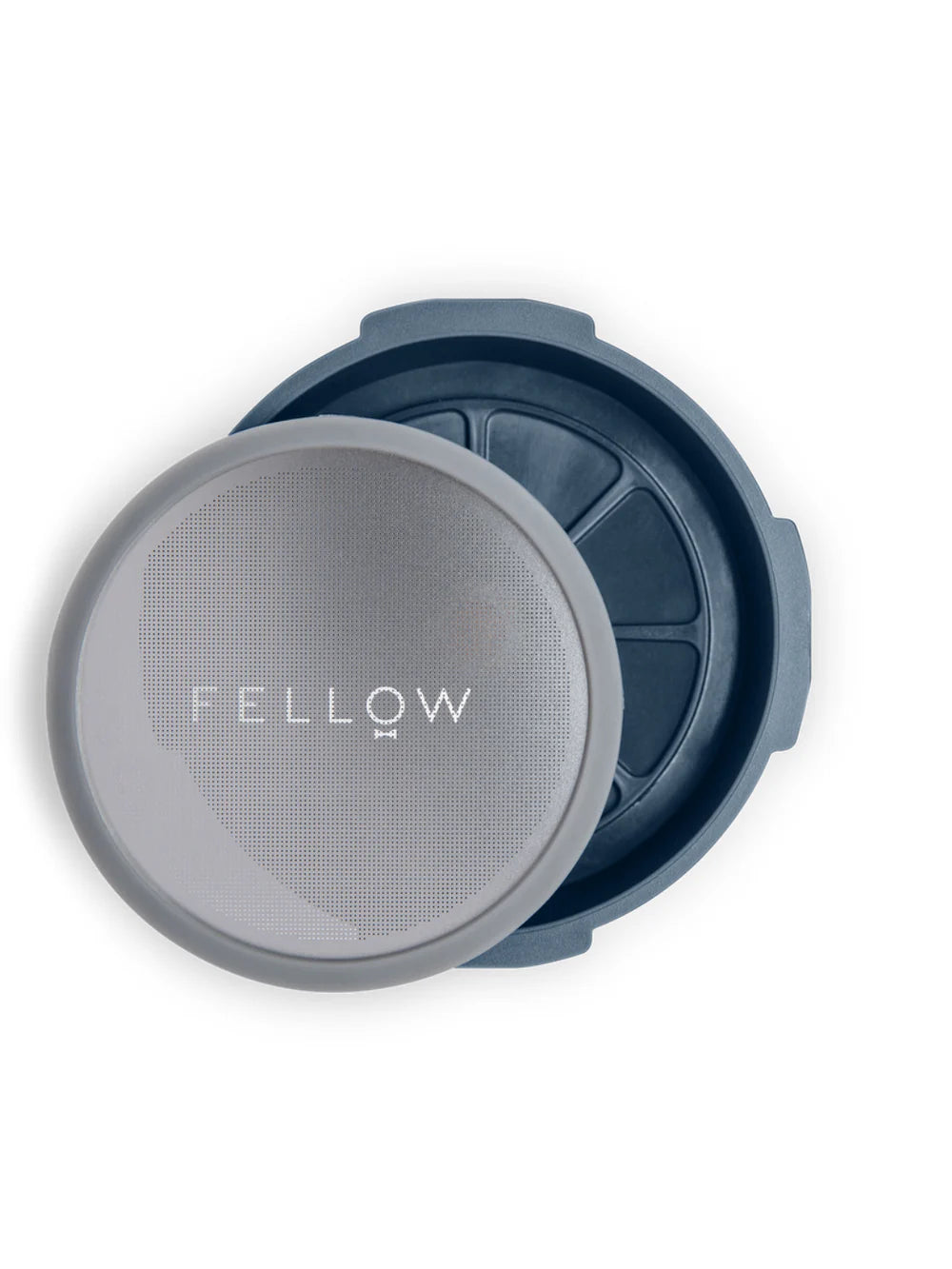 A close-up of a round gray mesh filter with the word 'FELLOW' printed on it, placed in front of a dark blue-gray container with segmented interior compartments, on a white background.